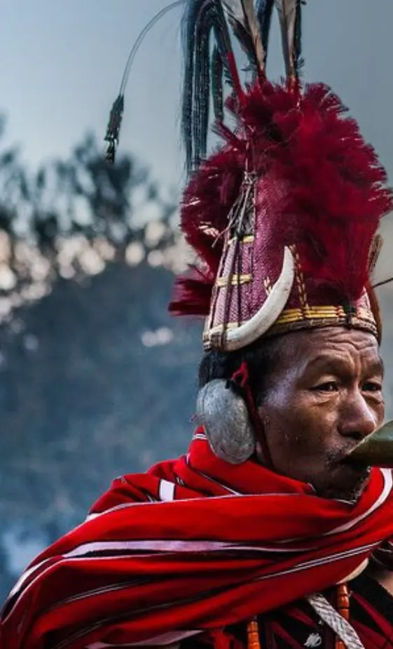 Naga elder in a red shawl and hornbill headdress blowing a horn